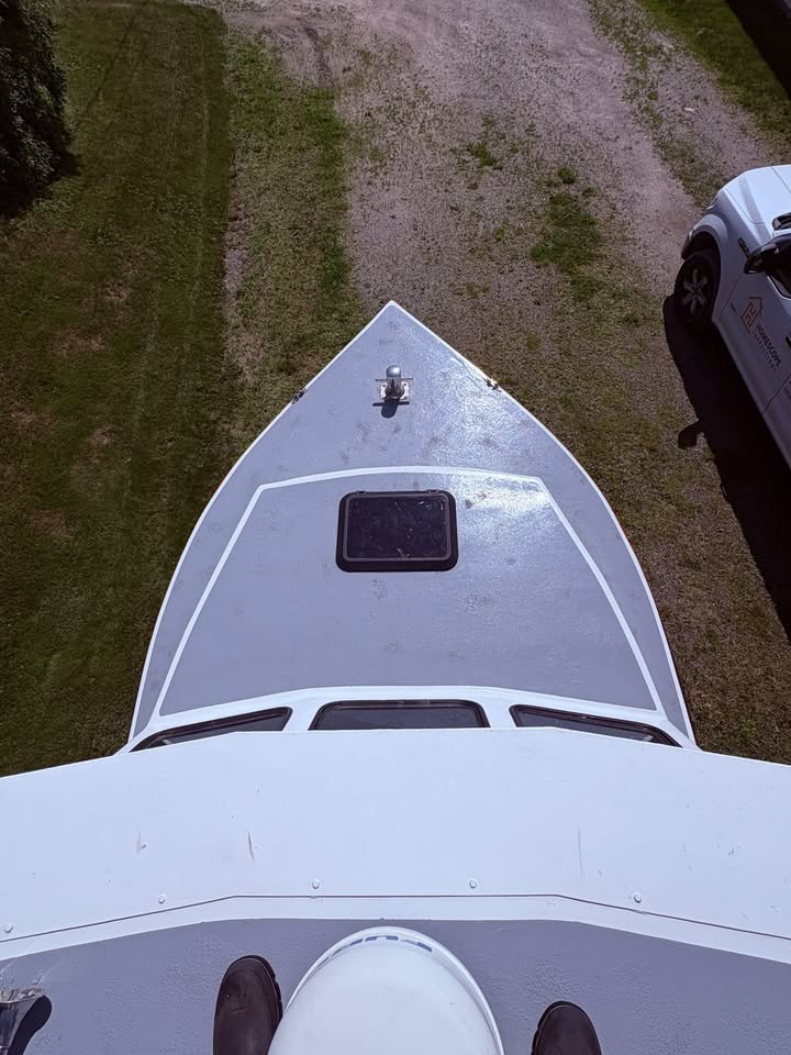 Aerial view from the bow of a white boat, showing the pointed hull, a small hatch window, and grass and dirt surrounding it. Part of a white vehicle is visible on the right.