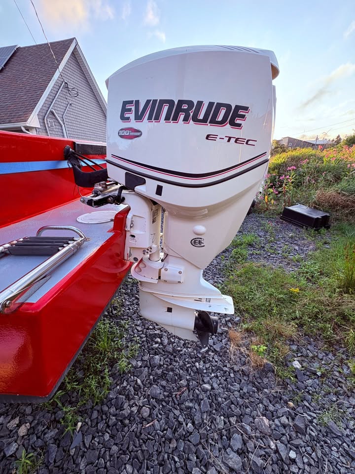 An Evinrude E-TEC outboard motor mounted on a red boat, photographed outdoors on a gravel surface next to a house.