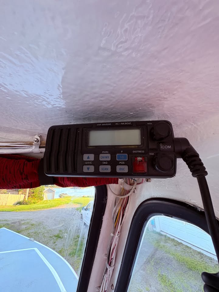 A black GCOM handheld marine radio transmitter mounted on the inside of a boat cabin window, with its power cord plugged in. The device features a digital display screen, blue and white buttons, and a red power switch.