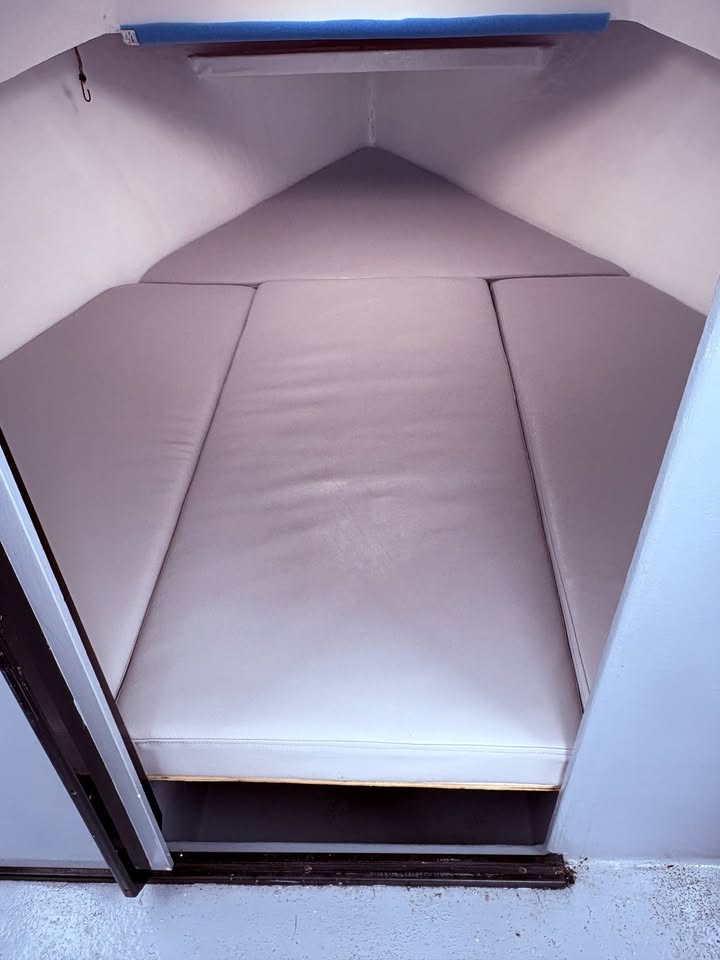 A close-up view of a compact bed or bunk-style sleeping berth with light beige padded mattress panels inside a narrow cabin or storage space.