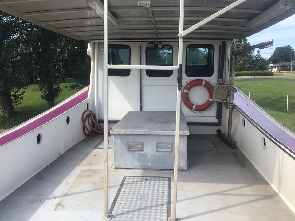 The back deck of a white boat or vessel with a gray metal storage box in the center, an orange life preserver, and a purple stripe along the side, photographed outdoors.