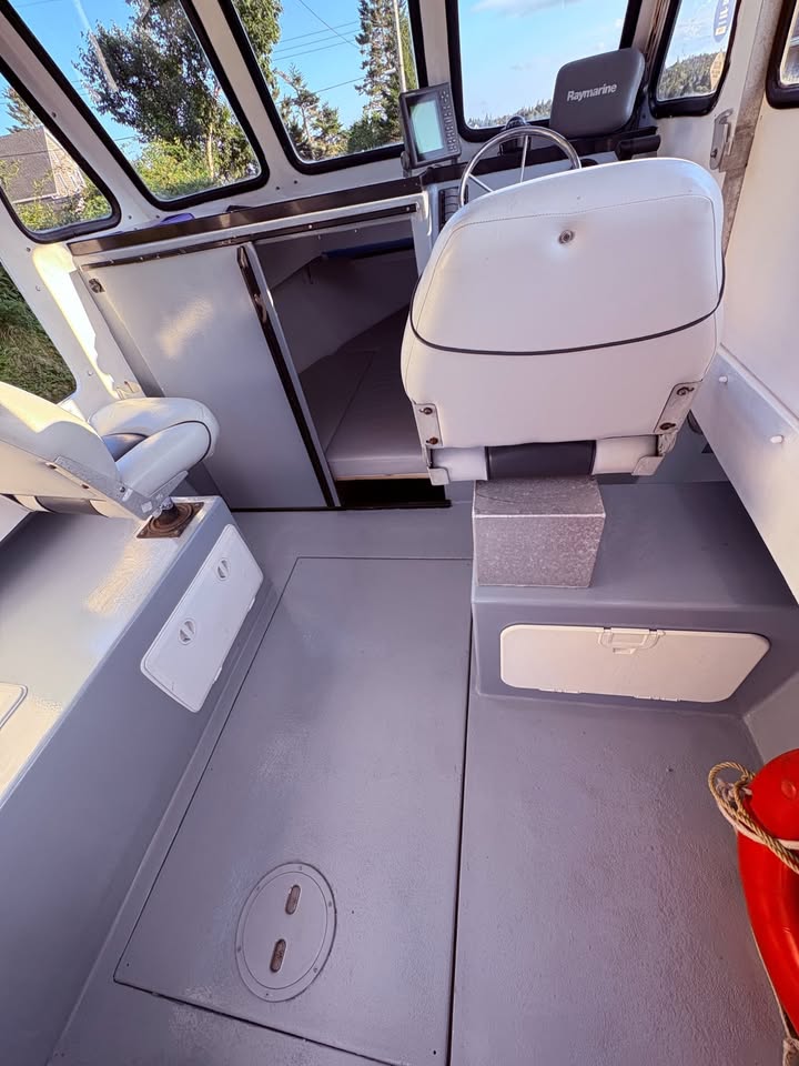 The interior of a boat cabin with gray flooring, white seats, a helm station with screens, and windows showing trees and sky outside.