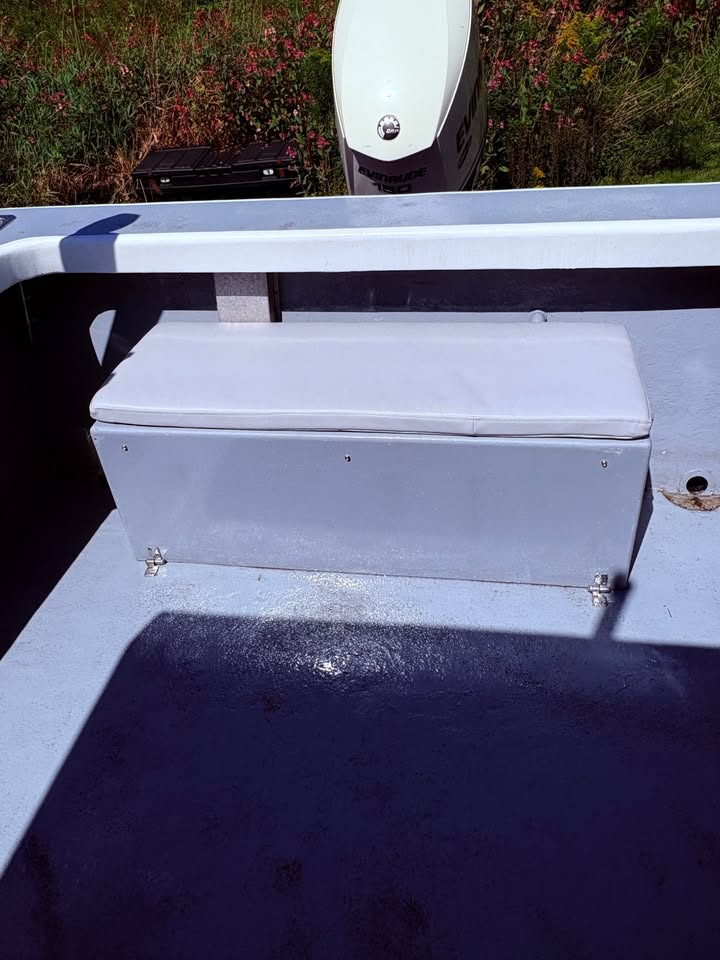 A light gray metal storage box with a matching lid, mounted on the deck of a boat. The boat is photographed outdoors on a sunny day, with a white outboard motor and green grass visible in the background.