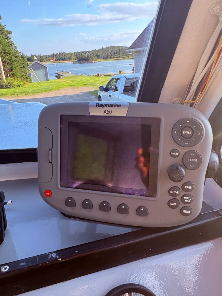 A Raymarine A60 fishfinder device resting on the dashboard inside a boat, with a scenic lake and shoreline visible through the window behind it.