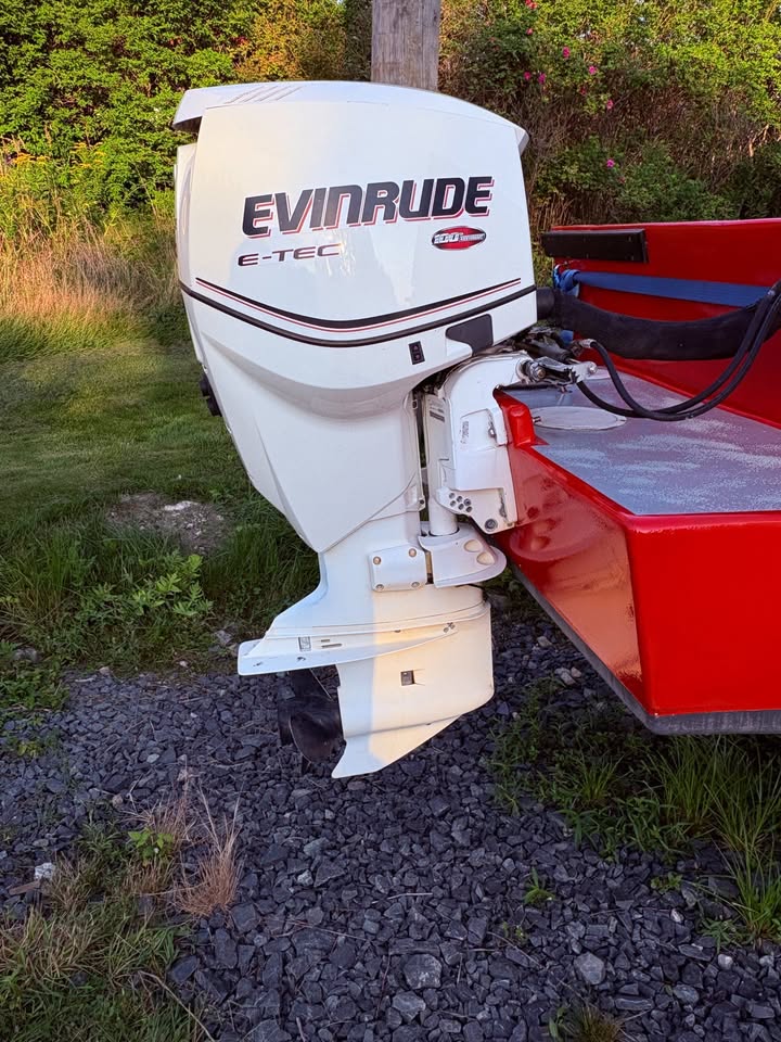 A white Evinrude E-TEC outboard motor mounted to the side of a red boat, photographed outdoors on a gravel surface with grass and trees in the background.
