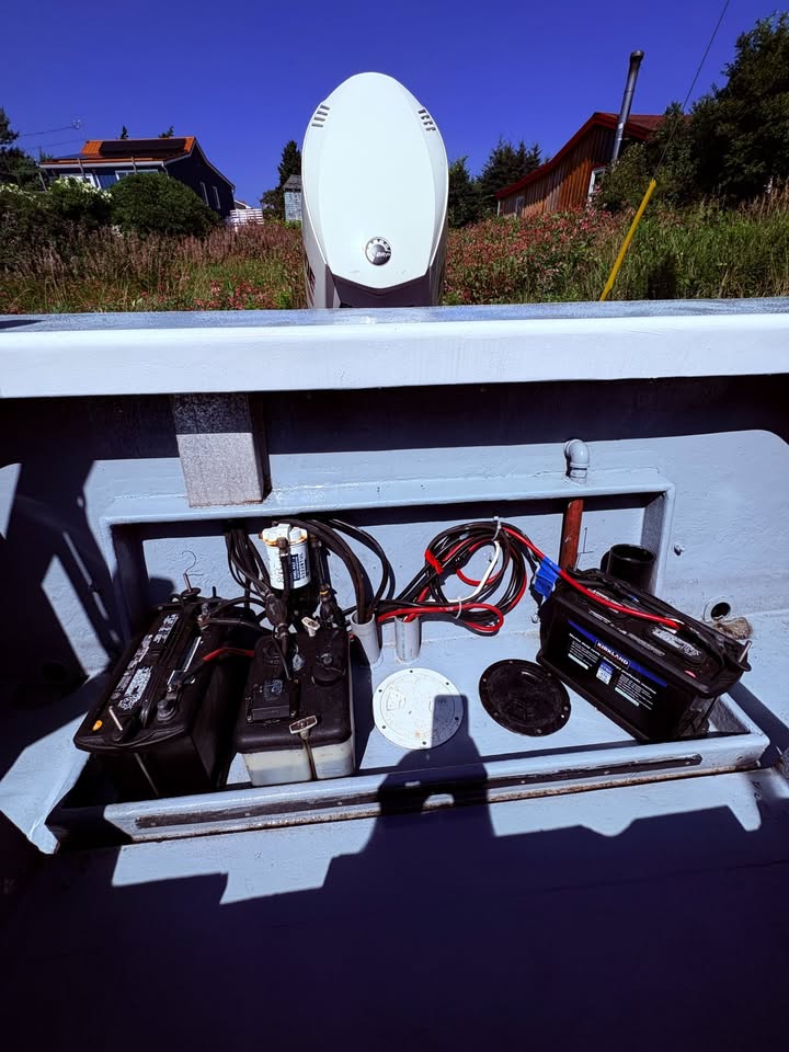 An open storage compartment of a boat showing various electrical components, including two black batteries with red and black cables, a white marine battery, and a circular white component, set against a blue sky and outdoor background.