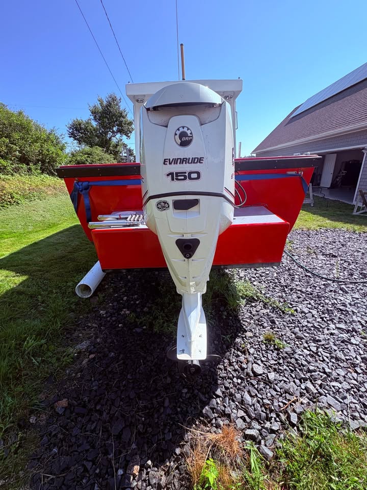 A red and white boat with an Evinrude 150 outboard motor parked outdoors on a gravel surface next to grass and a building under a blue sky.