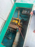 A close-up view of a boat engine compartment with a person's hand and foot visible, showing a yellow and black engine block, battery, wiring, and a large insulated pipe inside a teal painted engine bay.