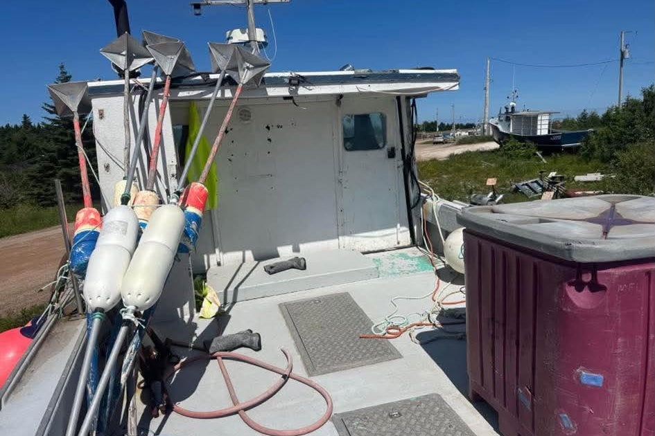 A narrow outdoor photo showing the deck and equipment of a small boat or vessel under a blue sky, with a large maroon container on the right and fishing-related gear on the left.