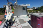 A narrow outdoor photo showing the deck and equipment of a small boat or vessel under a blue sky, with a large maroon container on the right and fishing-related gear on the left.