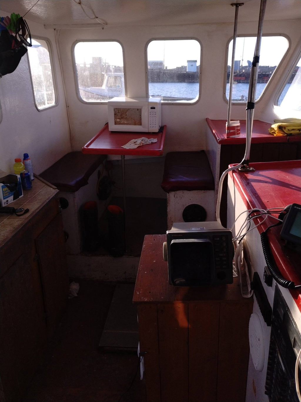 The interior of a boat cabin with red tables, dark cushioned seating, windows looking out onto water, and several electronic devices including a microwave and a small monitor on wooden consoles.
