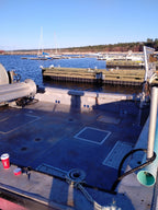 The back deck of a boat in a marina, featuring a blue non-slip deck mat, ropes, and a view of water, docks, and sailboats in the background under a clear blue sky.