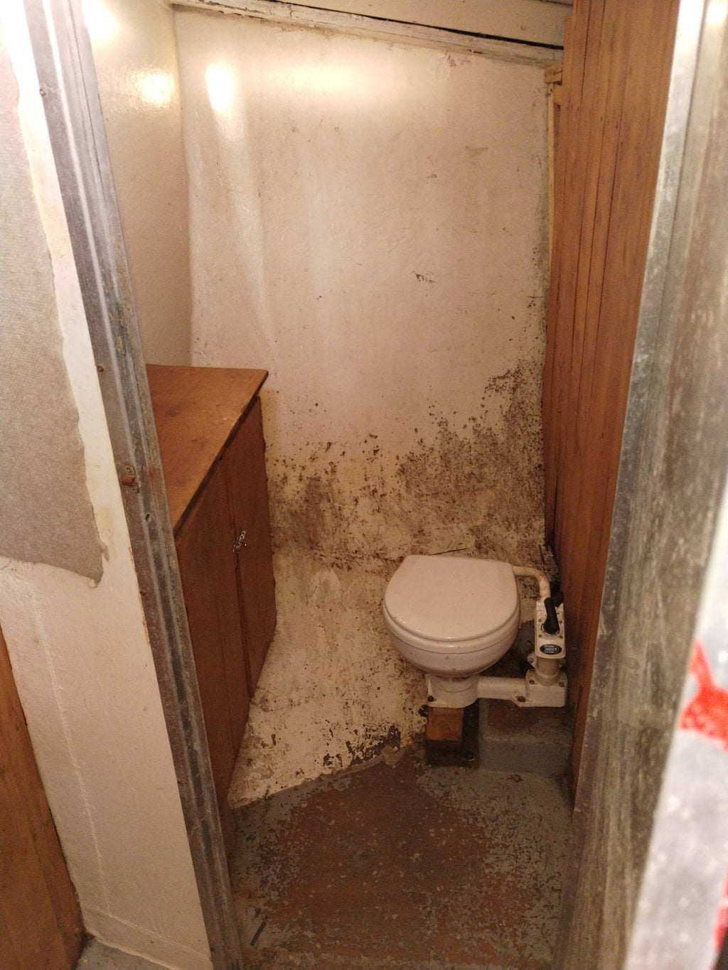 A small, dilapidated bathroom stall with a white toilet, a wooden vanity cabinet, and heavily stained beige and brown walls and floor.