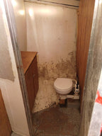 A small, dilapidated bathroom stall with a white toilet, a wooden vanity cabinet, and heavily stained beige and brown walls and floor.
