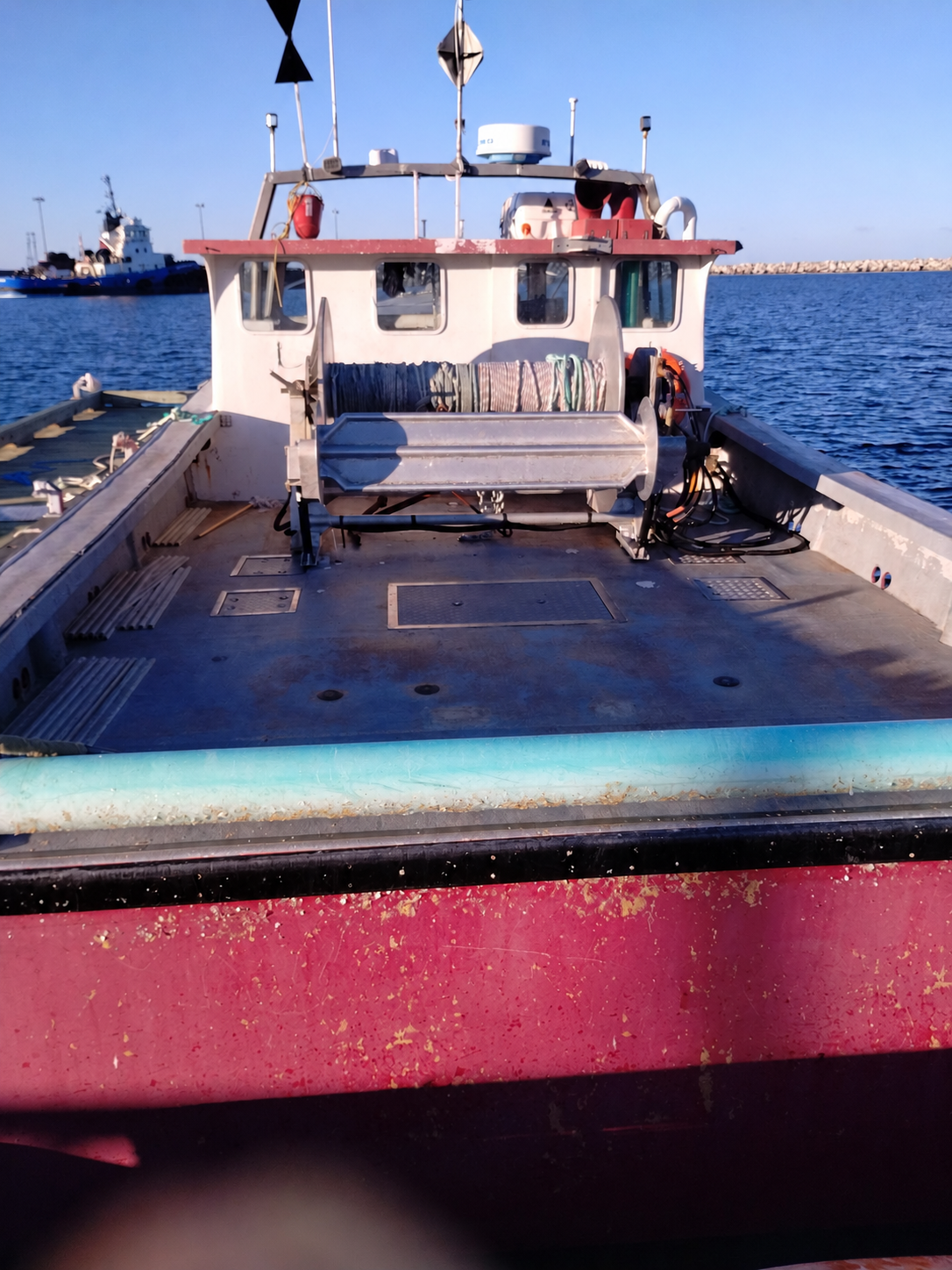 The back deck of a fishing boat on open water, featuring a gray bench seat, navigation equipment on the roof, and a weathered hull with a red upper section and a turquoise stripe.