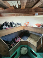 A rustic wooden loft bed with open storage compartments underneath, containing clothes and sports equipment, set against a white wall with exposed wooden ceiling beams.