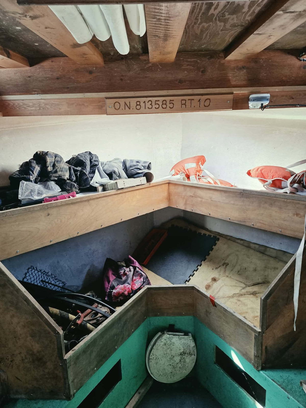 A rustic wooden loft bed with open storage compartments underneath, containing clothes and sports equipment, set against a white wall with exposed wooden ceiling beams.