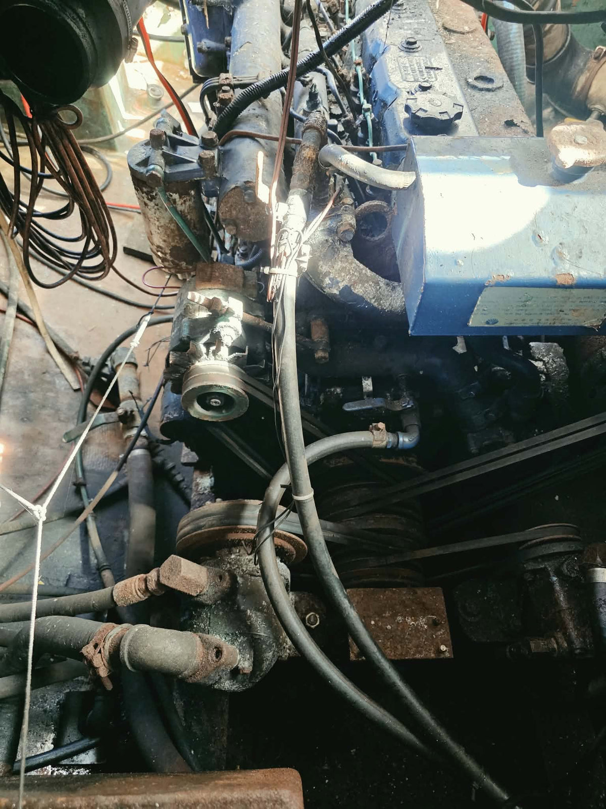 A close-up view of a used blue and black engine block with various hoses, belts, and metal components, photographed in a workshop setting.