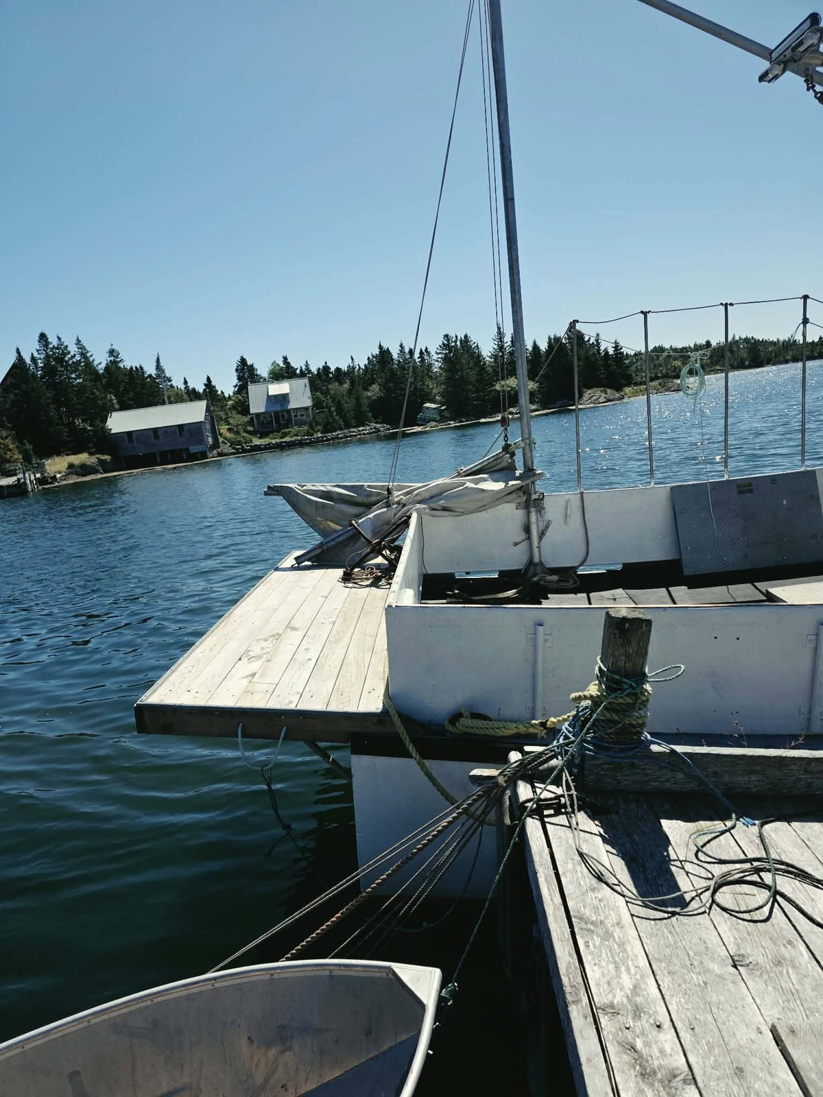 A close-up view from the deck of a white sailboat docked at a wooden pier, with blue water, a shoreline of trees, and small buildings in the background under a clear blue sky.
