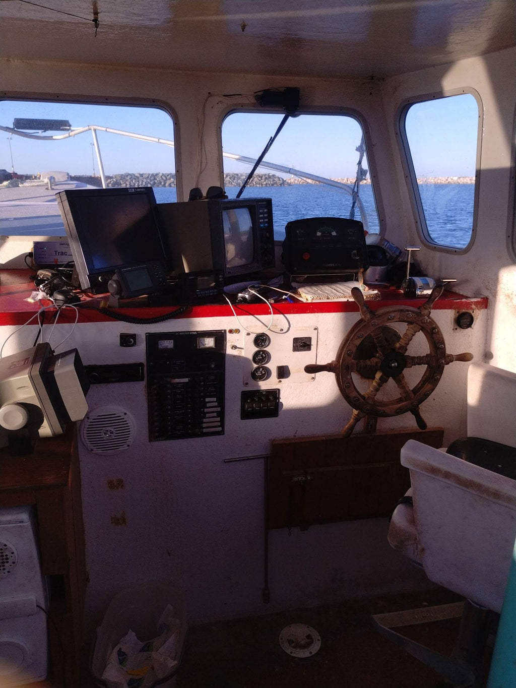 The interior of a boat cabin with a wooden ship's wheel, navigation screens, and control panels, with water and a bridge visible through the windows.