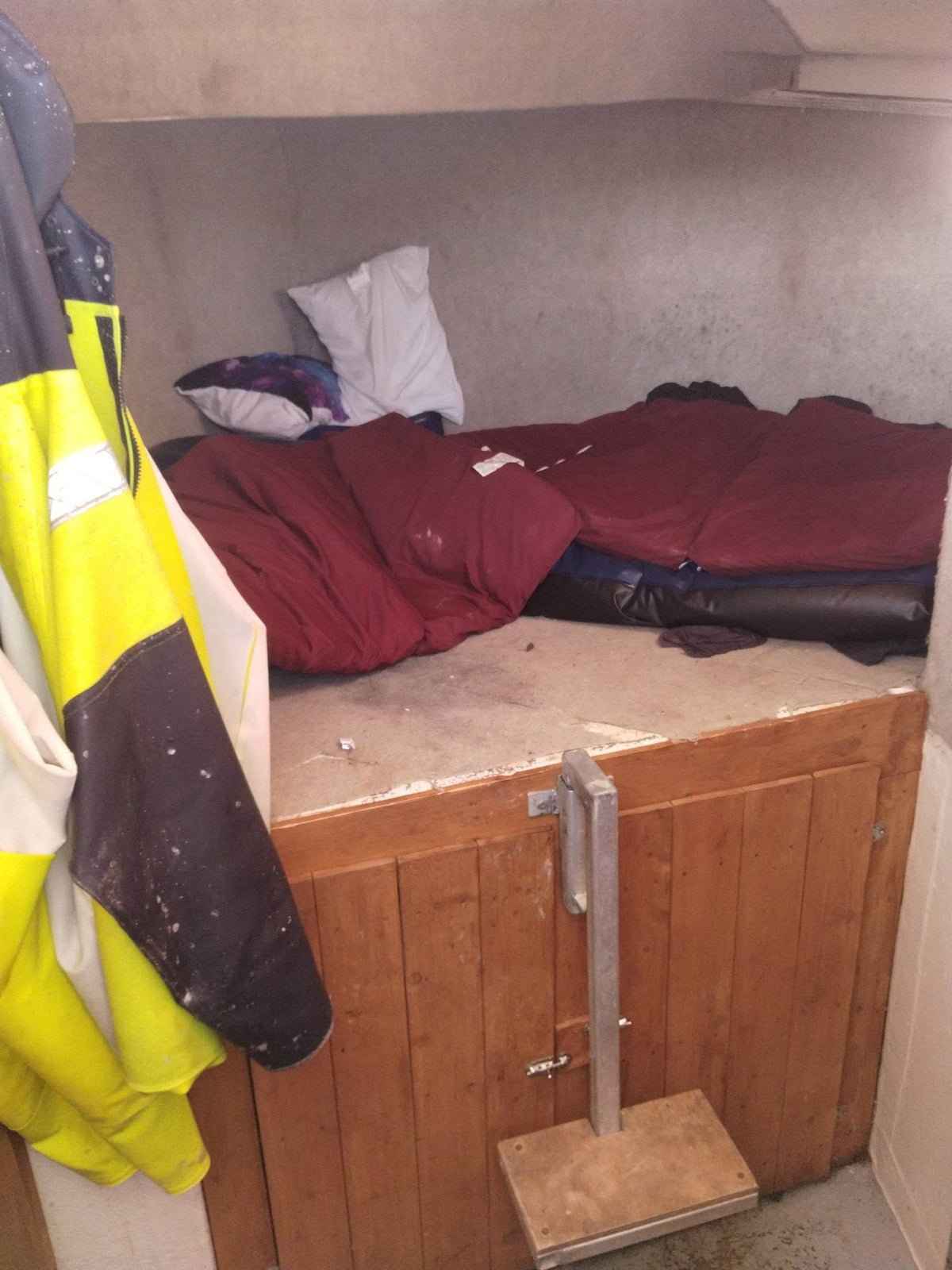 A cramped sleeping berth inside a boat or small cabin, with a wooden bunk bed, burgundy sleeping bag, white pillow, and a high-visibility yellow jacket hanging on the left.