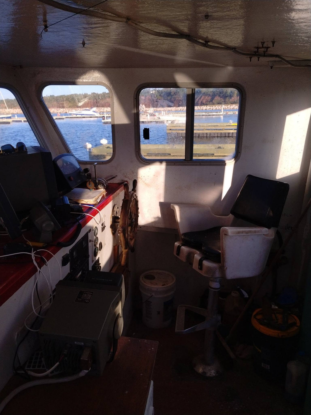 The interior of a boat cabin with a captain's chair, navigation console, windows showing water and docks outside, and various equipment on board.