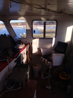 The interior of a boat cabin with a captain's chair, navigation console, windows showing water and docks outside, and various equipment on board.