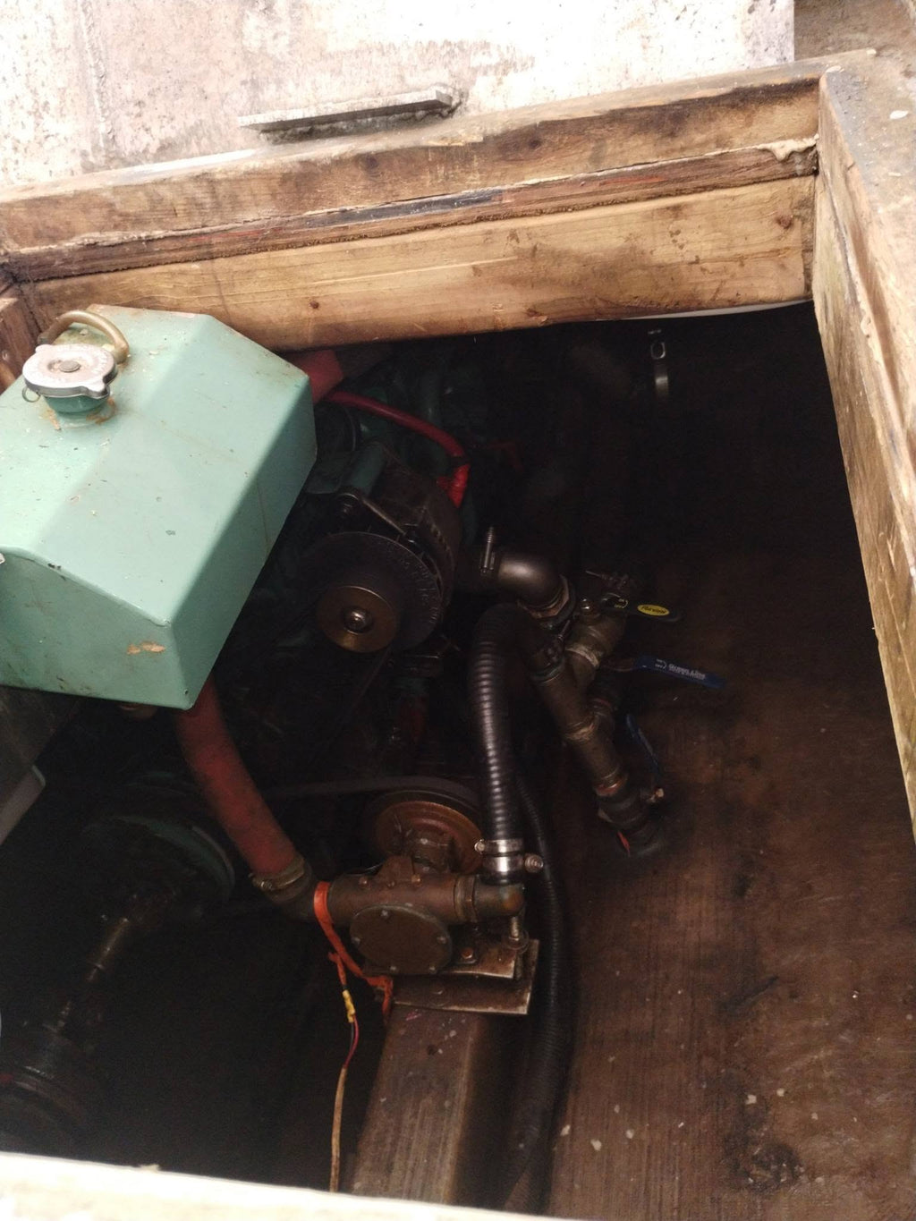 A close-up view of a boat engine or machinery compartment with a light green metal housing, black hoses, belts, and mechanical components inside a wooden opening.
