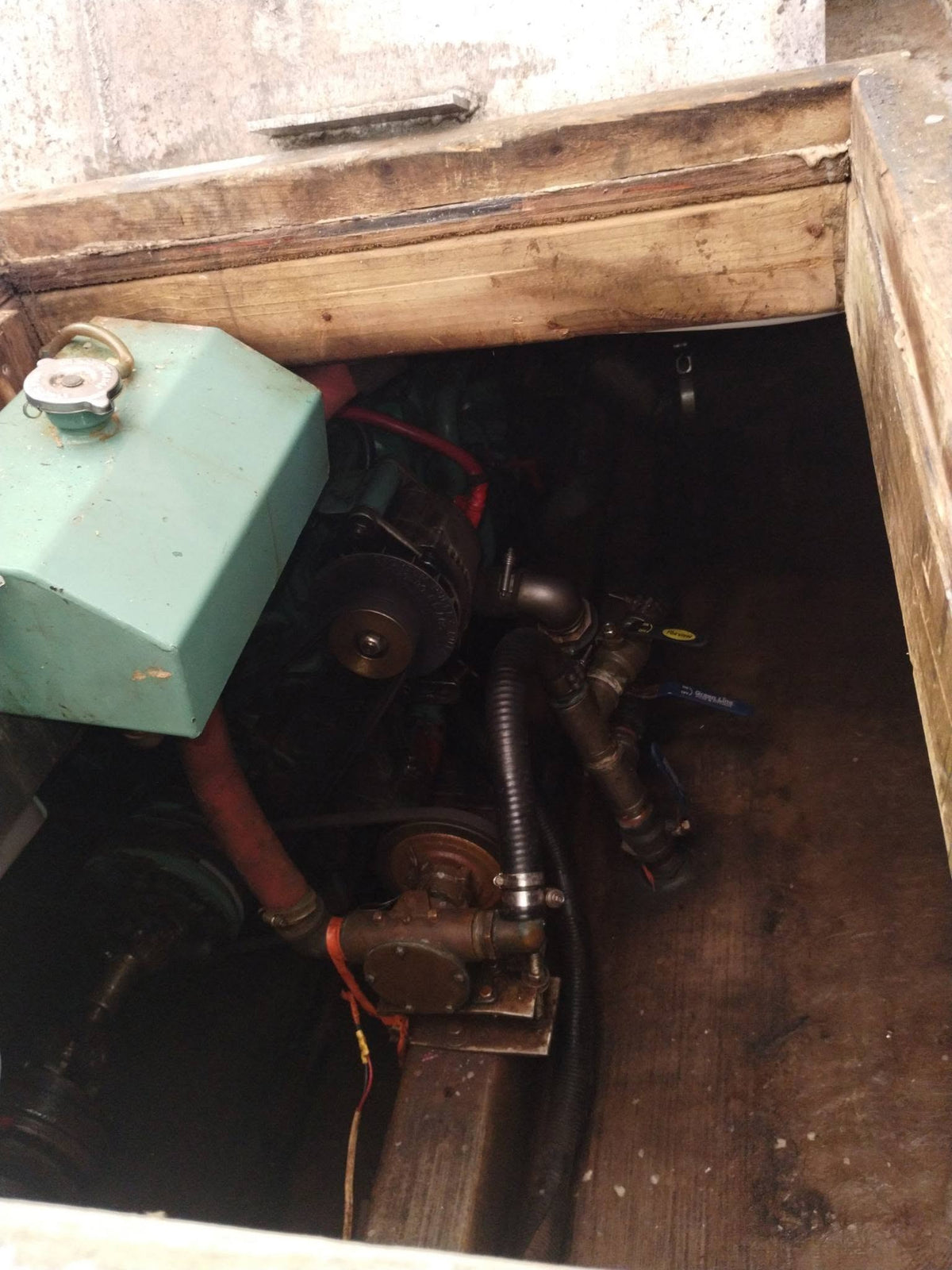 A close-up view of a boat engine or machinery compartment with a light green metal housing, black hoses, belts, and mechanical components inside a wooden opening.