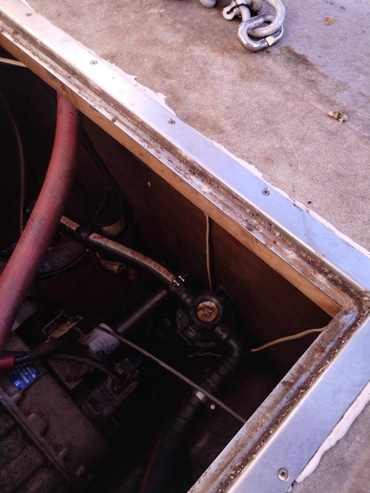 Close-up of a mechanical compartment or engine bay with hoses, pipes, wires, and a concrete floor, showing a red hose and dark metal components.