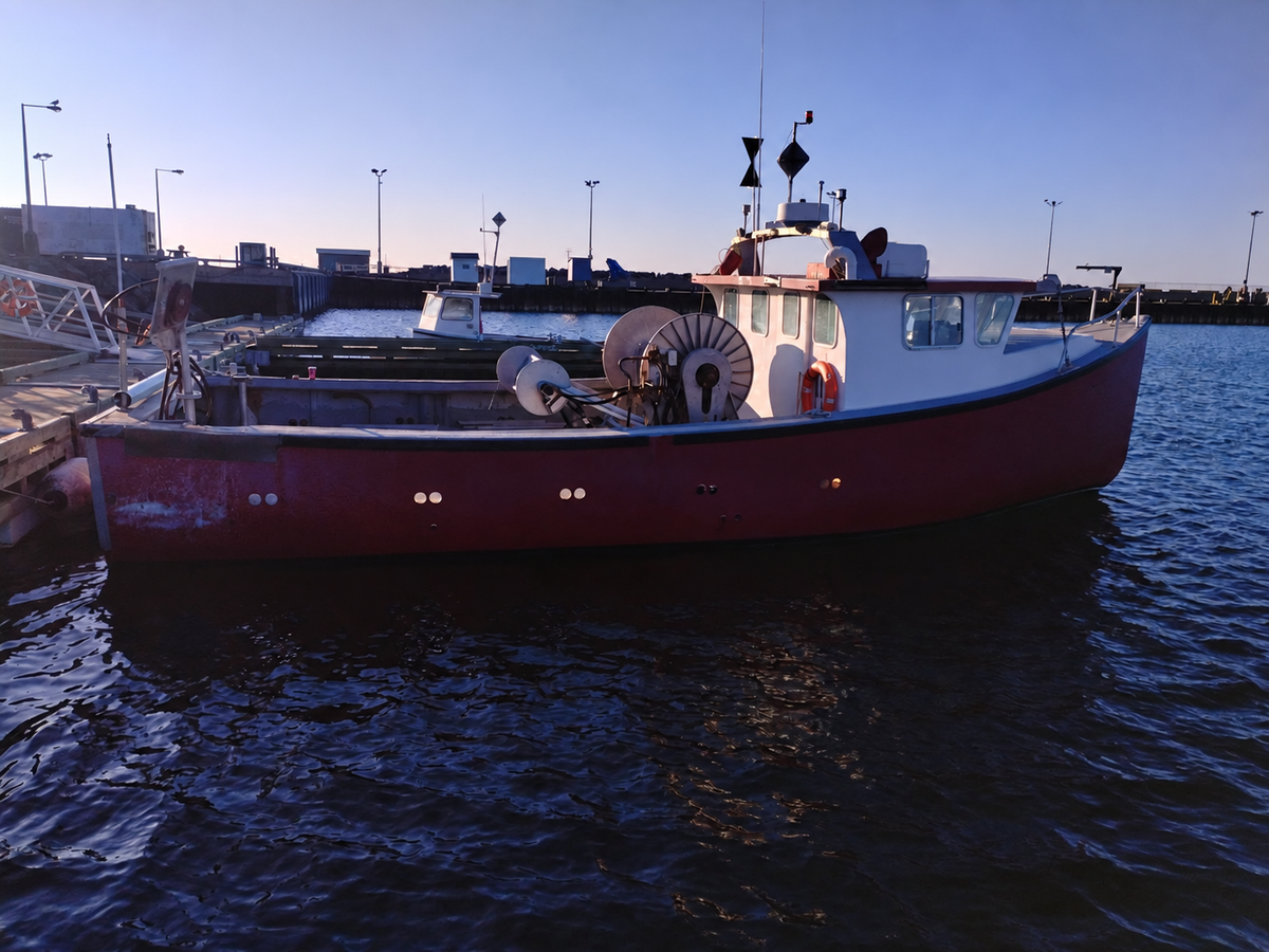 A red and white fishing boat docked at a harbor under a clear blue sky, with water and harbor structures visible in the background.