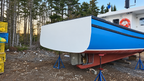 A blue and white boat hull resting on blue support stands on a gravel ground, with a forest background and a white vessel cabin visible behind it.