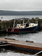 A classic wooden motorboat with a dark blue hull, reddish-brown deck, and cream cabin, docked at a wooden pier in calm water under an overcast sky.