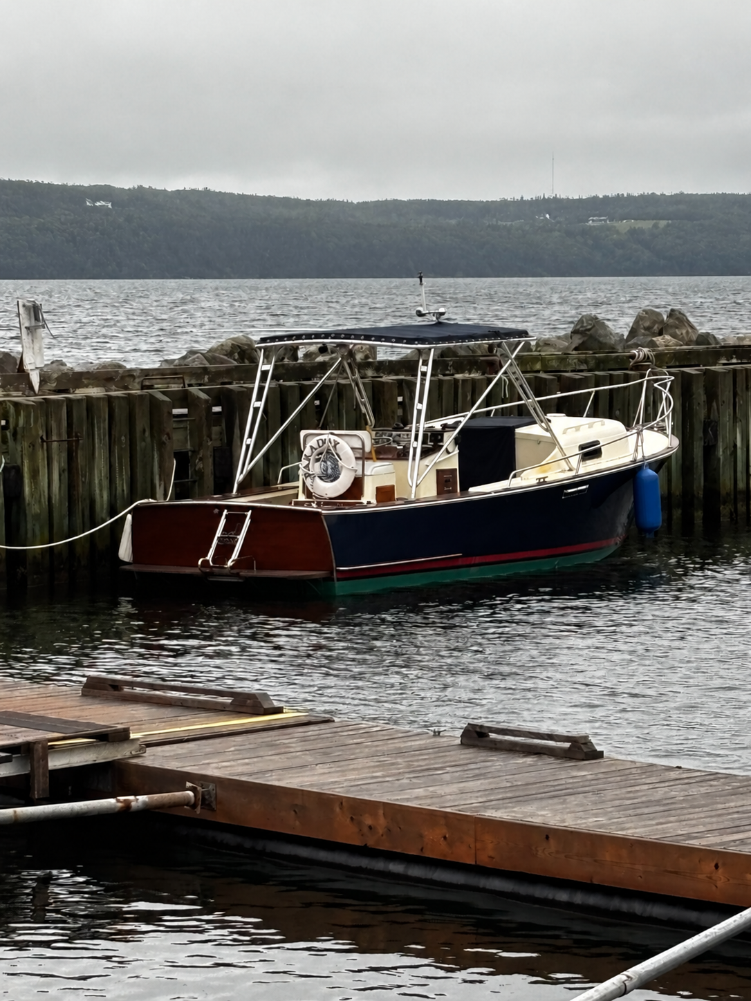 A classic wooden motorboat with a dark blue hull, reddish-brown deck, and cream cabin, docked at a wooden pier in calm water under an overcast sky.