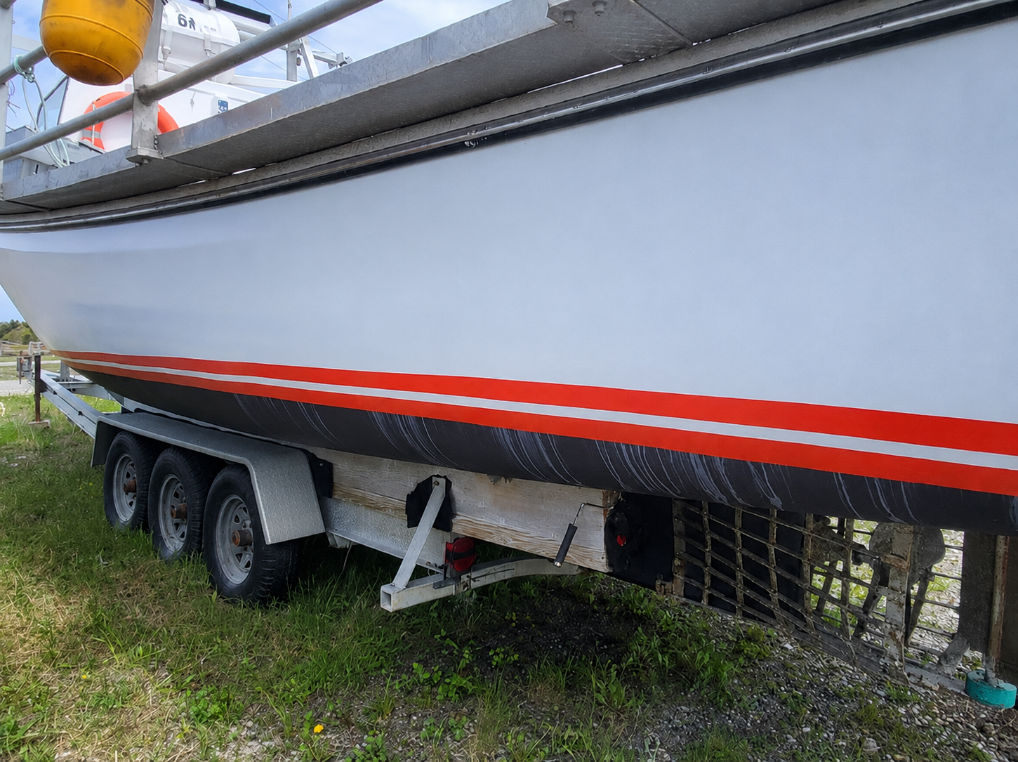 A white boat on a three-axle trailer, photographed outdoors on grass and gravel. The boat has a bold red stripe running horizontally along the hull.