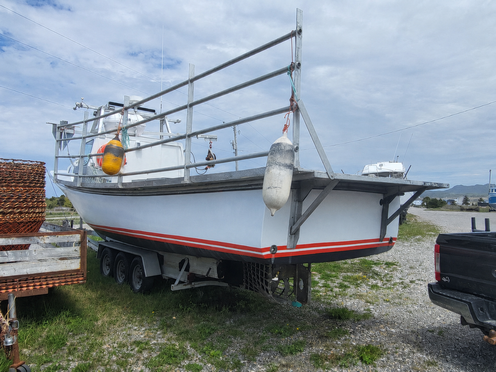 A white fishing boat on a trailer parked outdoors on a gravel lot under a cloudy sky, with metal railings, a yellow-and-white buoy, and part of a black pickup truck visible.
