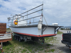 A white fishing boat on a trailer parked outdoors on a gravel lot under a cloudy sky, with metal railings, a yellow-and-white buoy, and part of a black pickup truck visible.