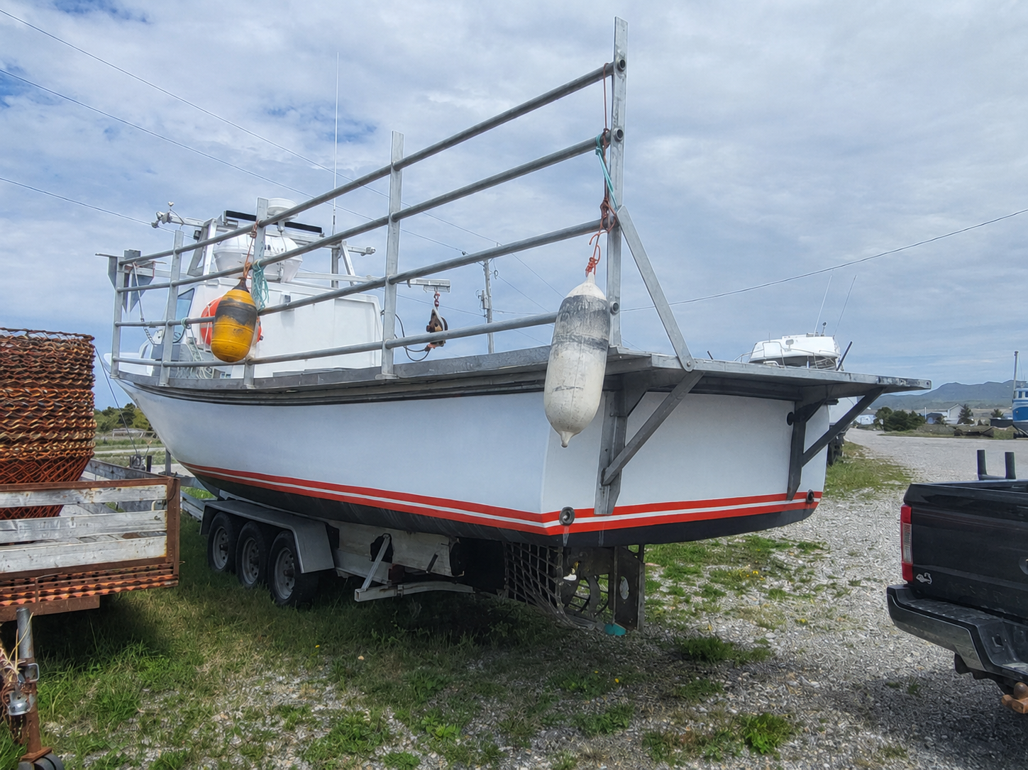 A white fishing boat on a trailer parked outdoors on a gravel lot under a cloudy sky, with metal railings, a yellow-and-white buoy, and part of a black pickup truck visible.