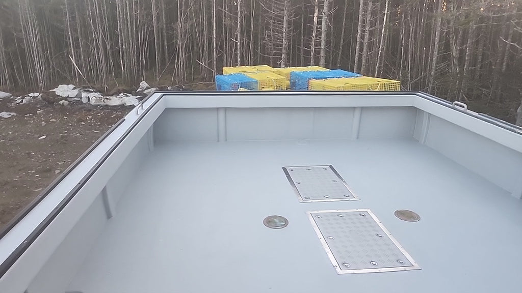 A light gray boat deck shown from above, with metal hatch covers and circular deck fixtures. Bare trees and a forest are visible in the background, along with yellow and blue bundled materials on the deck.