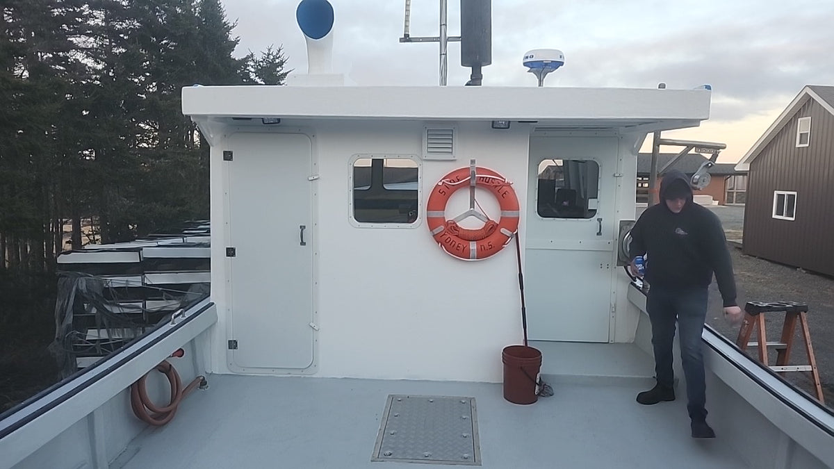 A man walks onto the deck of a white boat or vessel, with the cabin, orange life preserver, bucket, trees, and neighboring buildings visible in the background.