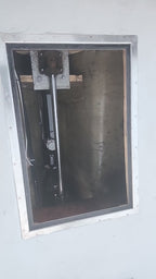 A close-up view of a metal-framed window or hatch opening, showing a dark interior with a vertical metal support and mounting hardware.