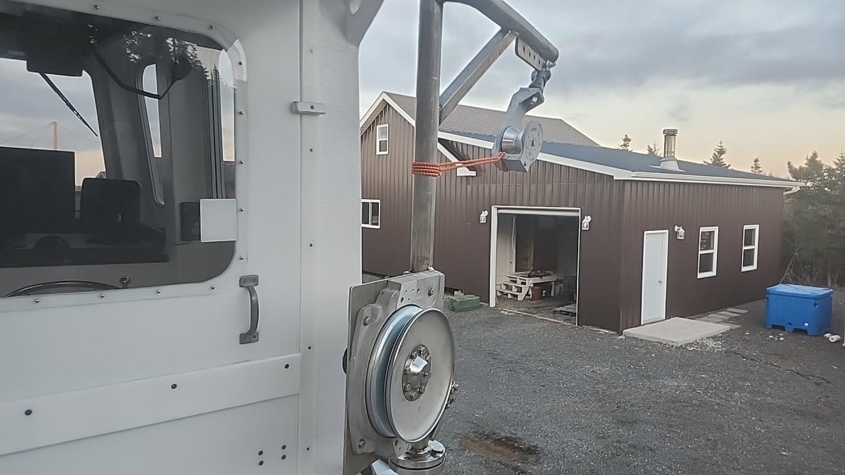 A close-up view of a white utility vehicle or machinery frame with a large pulley system and metal guide rod, set outdoors in front of a brown metal workshop building and an overcast sky.