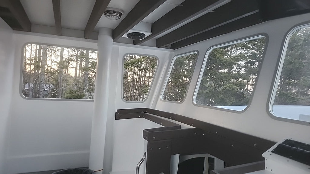 The interior of a boat cabin with white walls and windows, dark wood trim, and a view of trees outside.