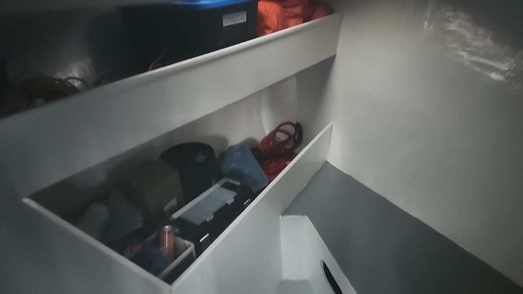 A low-angle view inside a white storage compartment or cabin interior, showing shelves with various items including a black box, orange fabric, and other miscellaneous objects.