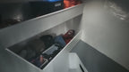 A low-angle view inside a white storage compartment or cabin interior, showing shelves with various items including a black box, orange fabric, and other miscellaneous objects.