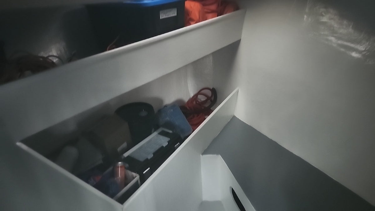 A low-angle view inside a white storage compartment or cabin interior, showing shelves with various items including a black box, orange fabric, and other miscellaneous objects.