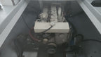 A close-up view of a boat's engine housed within a gray fiberglass compartment, showing the engine block, belts, and wiring.