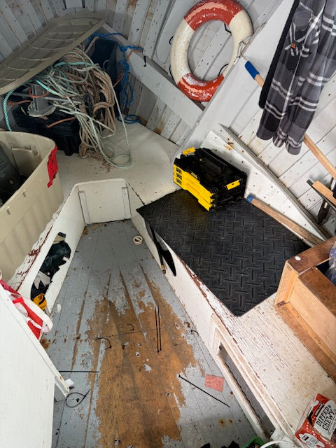 The interior of a weathered white boat or cabin with a worn wooden floor, a black diamond-plate mat, a yellow and black toolbox, a red and white life preserver, and various storage items in the background.