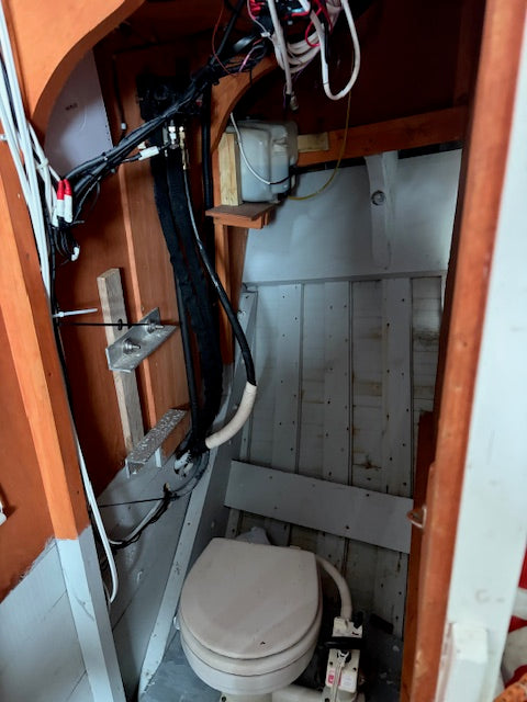 A compact, utilitarian bathroom or toilet cubby with exposed wiring, wooden framing, and a white toilet seat, likely installed in a boat or small vessel.