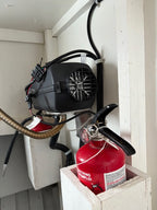 A red fire extinguisher mounted in a white wooden box on a wall, with a black portable air pump or compressor attached above it and hoses connected.
